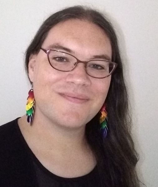 A picture of myself, a white trans woman with long dark hair over one shoulder and rainbow earrings